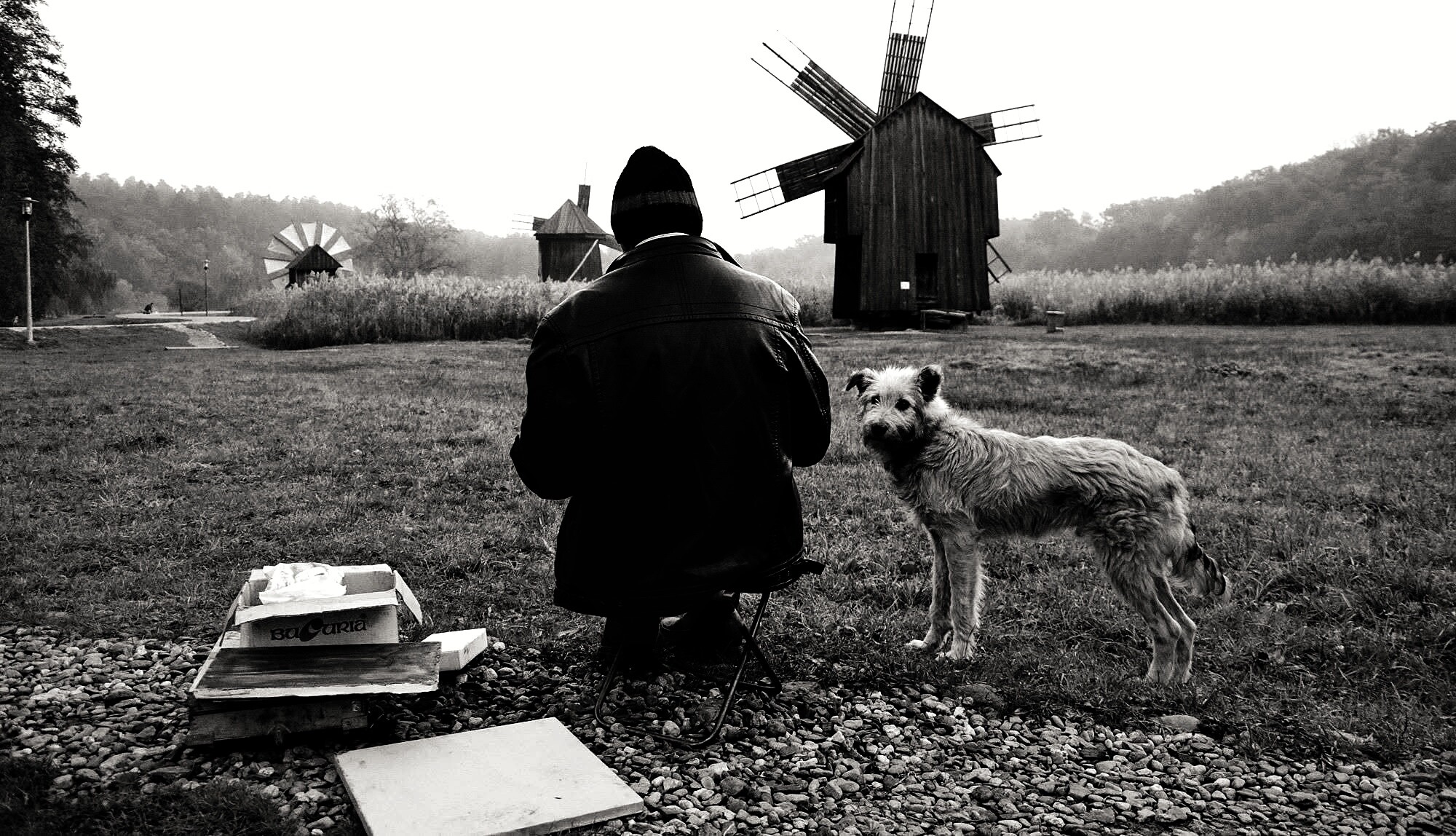 Archival photograph, 2015, Sibiu, Romania; person working outdoors during an educational plein-air art programme, with a dog and wooden windmills in the landscape.